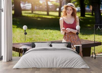 Photo of attractive woman with foxy hair, wearing hat, floor skirt and maroon t shirt, sitting on bench in city park, charging smart phone via alternative energy. Alternative energy, ecology concept. Wall mural