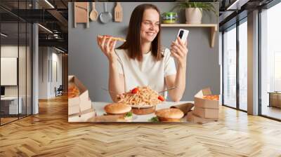 Photo of cheerful Caucasian young adult woman with brown hair wearing white t shirt sitting in kitchen at table and looking at smart phone display, having delicious snack, eating pizza. Wall mural