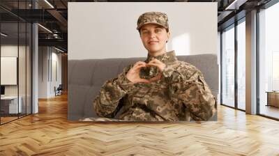 portrait of of caucasian smiling soldier female wearing camouflage uniform sitting on sofa and looki Wall mural