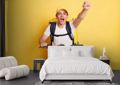 Portrait of positive happy optimistic deliveryman on bicycle wearing white T-shirt and cap isolated over yellow background, raised fist, making announcement. Wall mural