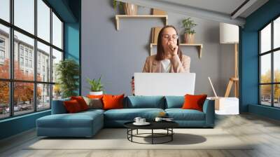 Portrait of sleepless tired woman office manager with dark hair sitting at table and working on laptop, being exhausted, yawning, covering mouth with hand, being sleepy. Wall mural