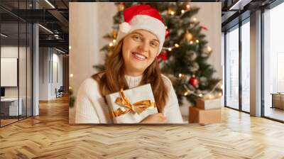 Portrait of smiling woman holding New Year gift in box with gold ribbon, happy female wearing santa hat and white sweater posing indoor with Christmas tree on background. Wall mural