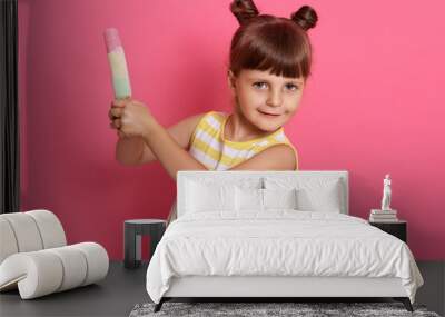 Satisfied little child holding water ice cream with both hands, looks at camera, has two knots, wearing striped summer dress, posing isolated over rosy background. Wall mural