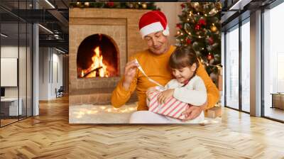 Senior man with his granddaughter sitting on floor and opening present box, old male in yellow shirt and red festive hat helps to little girl to see her present, posing with xmas tree on background. Wall mural