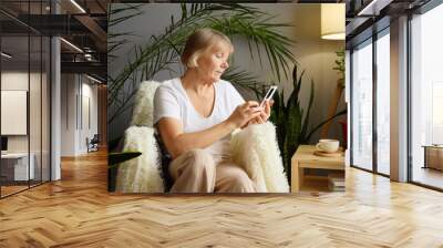 Senior woman holding smartphone browsing internet while scrolling through online content in comfort of her home using modern technology to read and watch digital media Wall mural