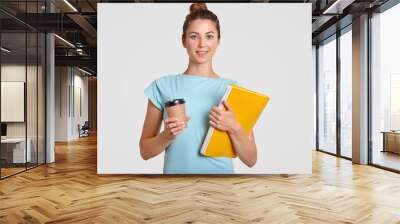 Shot of pleasant looking woman with hair bun has delighted expression, holds takeaway coffee and yellow folder, dressed casually, rests after having classes, isolated over white studio background Wall mural