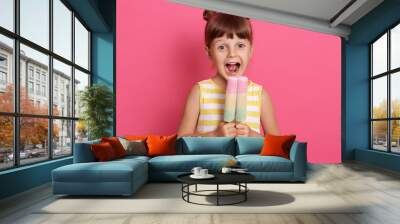 Small European kid biting fruit ice cream, little charming girl with widely opened mouth, wearing summer clothes, looks happy, having fun with delicious dessert. Wall mural