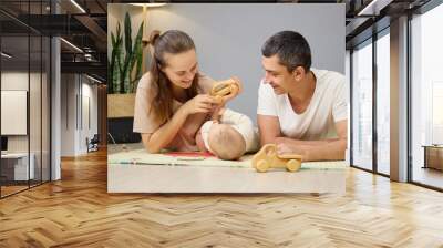 Smiling cheerful woman and man lying on floor with infant interacting with toddler child together enjoying babyhood moments of care and development in nursery room at home Wall mural