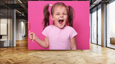 Studio shot of little attractive child opening mouth widely, looking directly at camera with excitement, holding bright lollipop in hand, wearing casually, isolated over pink studio background. Wall mural