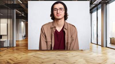 Waist up studio shot of delighted calm guy standing straight, looking directly at camera, wearing beige jacket, red sweatshirt and round trendy eyeglasses, posing isolated over white background. Wall mural