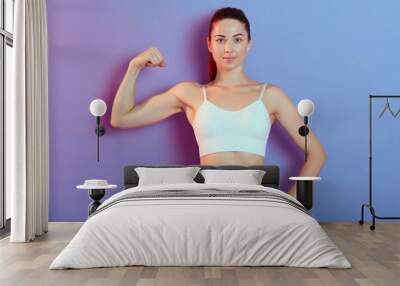 Winsome young woman showing off muscles, keep one hand on hip, looks at camera with confident facial expression, wearing white tank top and grey leggins, poses against color wall. Wall mural