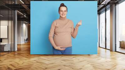 Young beautiful pregnant woman expecting baby, posing isolated on blue background, making korean like sign, doing positive gesture, being happy, cheerful expression, wearing casual clothing. Wall mural