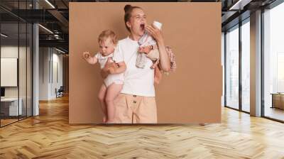 Young mother with dark hair wearing white t shirt standing with her baby daughter in hands, yawning, needs more sleep, having sleepless nights during maternity leave, isolated over brown background. Wall mural