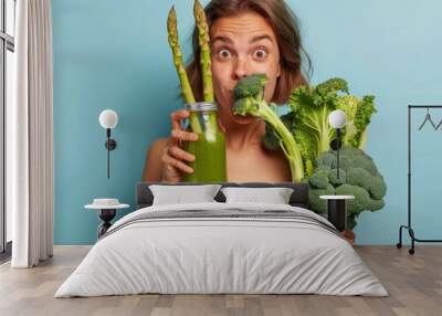 Woman holding various green vegetables and a mason jar with green smoothie against a blue background. Wall mural