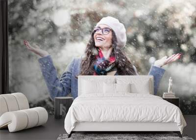 Portrait shot of attractive young woman wearing hat and winter coat while standing outside and enjoy falling snow. Happy woman standing outdoor and raising her hands while snowing heavily  Wall mural