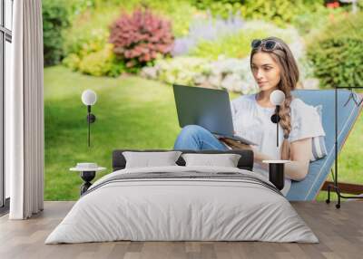 Smiling young woman using her laptop while sitting in the backyard at home Wall mural