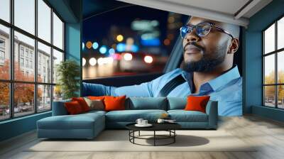 A stylish black man in glasses travels home in the back seat of a taxi at night. A handsome male passenger glances out the window of his car on an urban street with neon signs and a working traffic Wall mural