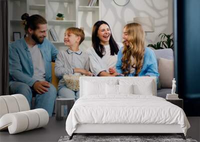 Attractive happy smiling parents and their two kids eating popcorn while sitting on the couch in the living room and watching TV. Cheerful mother, father, son and daughter spend time together at home. Wall mural