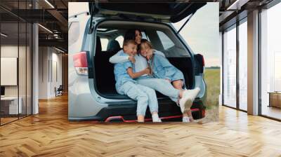 Charming loving joyful mom hugging her cheerful teen son and daughter while they together sitting in auto trunk,stopped in the green field during travel countryside,family concept Wall mural