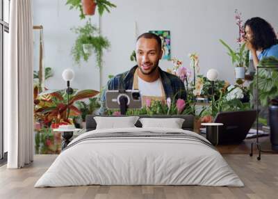 Handsome man gardener in apron using phone and recording blog video with clients. Young adult content creator influencer blogger recording a video talking about indoor plants care from workplace. Wall mural