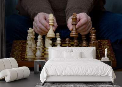 Hands of a white-skinned man holding two chess pieces over a game board showing them to the camera. Wall mural