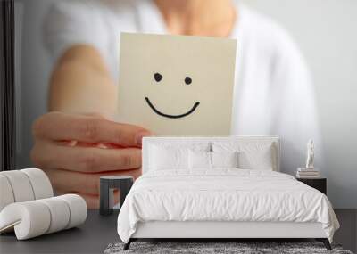 a young woman holds a paper sticker with a drawn smiley face in her outstretched hand. Focus on the hand and sticker, blurred background Wall mural