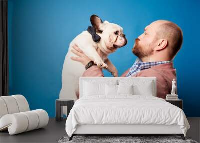 Bald man in jacket with french bulldog in his arms on empty blue background Wall mural