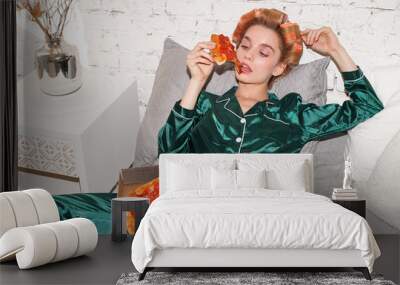 young fashionable woman in silk pajamas lays on bed and eat pizza pepperoni from pizza box, direct flash Wall mural