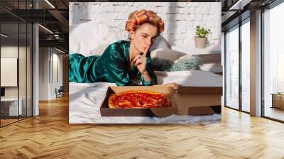 young woman in silk pajama showing silence gesture with pizza pepperoni in bed Wall mural