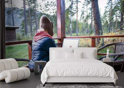 Female blogger concentrated on keyboarding on netbook article for web page sitting at table on terrace of house in autumn forest. Wall mural