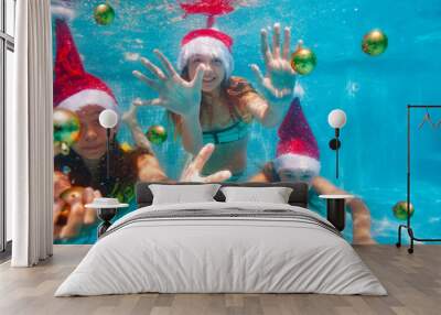 Three happy girls dive and swim underwater catching Christmas tree balls wearing Santa Claus hat in the pool Wall mural