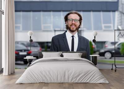 Portrait of a young businessman with a beard and long hair on the background of a business center with cars Wall mural