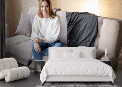 smiling young girl in a white sweater and jeans sits on a sofa in a cozy room and looks at the camera Wall mural