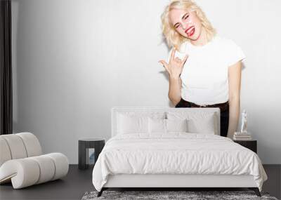 young stylish blonde girl in a white t-shirt and black jeans shows a rock sign on a white background, studio fashion photo with copy space Wall mural