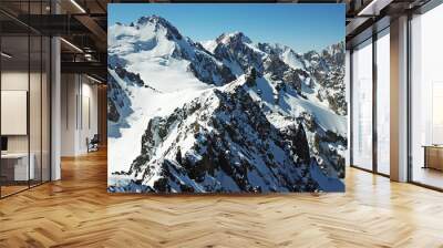 Snow-capped mountains and blue sky. View from a drone. Huge peaks covered with snow, steep cliffs. Blue, clear sky above the mountains. Flying over the peaks. Below you can see green trees and tarva. Wall mural