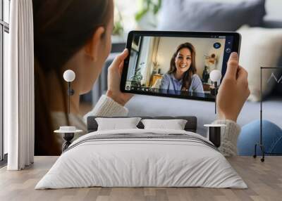 A woman is holding a tablet and engaging in a video call while sitting in a cozy indoor setting, capturing a relaxed moment. Wall mural