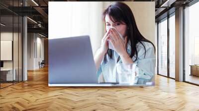 A young sick woman blows her nose at a laptop in the office - A girl is ill and wipes her nose with a napkin - An unhealthy employee suffers from an allergic reaction or a cold and flu - Sick person Wall mural