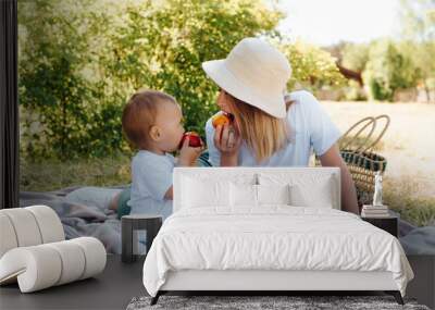 Family picnic, mom and baby eat fruit together, outdoors. Young mother in a hat and little son are resting on a blanket on the green grass outside, summertime. Family lifestyle Wall mural