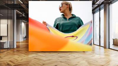 Fashionable young woman with glasses and red lipstick holds rainbow flag, international symbol of LGBTQ+ community, gay, lesbian, people of color and transperson. Equal rights and freedoms for LGBT Wall mural