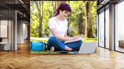 Focused student with purple hair studying outdoors with a laptop and notebook in a park. Education concept Wall mural