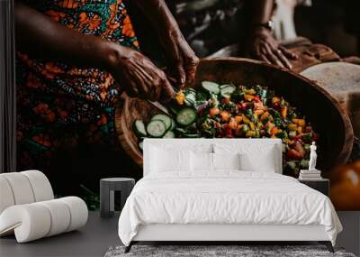 Hands of an elderly Black woman preparing a colorful fresh salad with cucumbers, bell peppers, and other vegetables, highlighting the details of preparation in a rustic and homely setting Wall mural