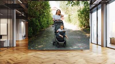 Happy young mother walking with her kid in stroller together in park on sunny summer day. Family, motherhood, parenting concept Wall mural