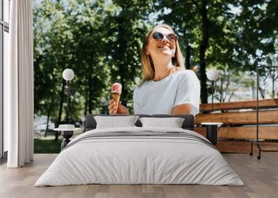 Joyful young woman relaxing on a park bench looking up enjoying the sun and summer weather. Cute smiling girl eating ice cream in a waffle cone, outdoors. Summer lifestyle Wall mural