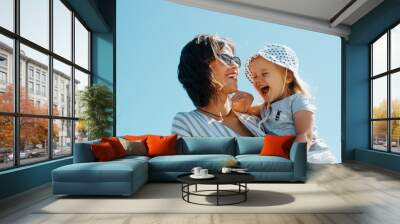 Positive family portrait laughing mom and daughter outdoors. Cheerful young beautiful mother holding laughing baby in her arms against blue sky Wall mural