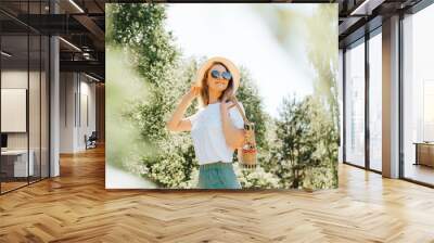 Pretty smiling caucasian young woman with freckles in straw hat, glasses and handbag looking away while standing in nature on sunny summer day. Portrait of happy charming girl walking outdoors Wall mural