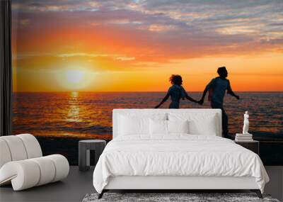 Silhouette of happy young couple in love together holding hands and running along seashore at sunset, copy space. Romance, relationship, date, valentine's day concept Wall mural