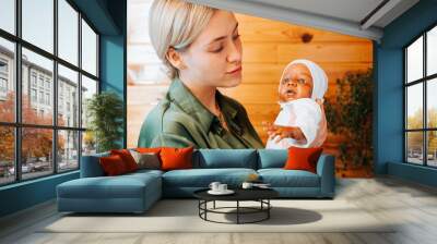 Smiling caucasian young woman holding newborn african american baby doll in arms and looking at her, indoors. Toy child reborn handmade Wall mural