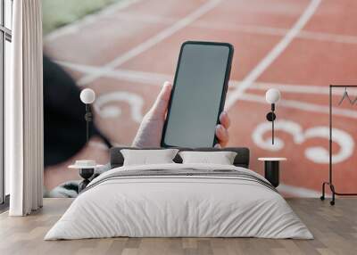 Sports and technology concept. Sportswoman using mobile phone with blank white screen while exercising in outdoor stadium. Close-up of woman's hand holding smartphone with an empty advertising space Wall mural