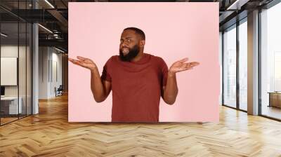 Handsome confused African-American model in stylish brown t-shirt shrugs standing on light pink background in studio closeup Wall mural