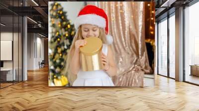 Little cute girl in Santa hat opens a Christmas present. Wall mural